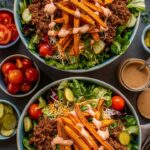 Two large, rustic bowls showcase loaded salad bowls filled with crisp green lettuce, seasoned ground beef, shredded cheddar cheese, diced ripe tomatoes, and tangy pickle slices. Oven-roasted sweet potato fries rest atop the greens, all generously drizzled with a creamy, light-brown dressing. Surrounding the main bowls are small dishes of extra chopped tomatoes, pickles, and sweet potato fries, plus a glass jar of dressing, fork, and linen napkin—creating a vibrant, farm-to-table presentation ideal for a hearty, nutritious meal.