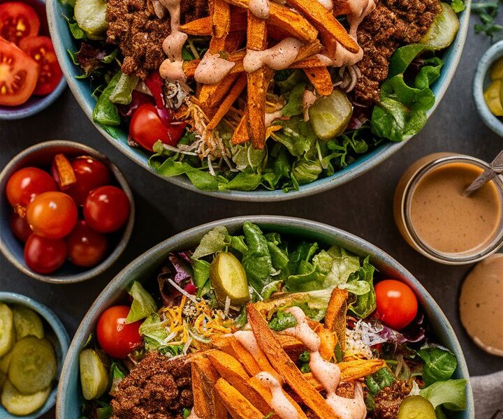 Two large, rustic bowls showcase loaded salad bowls filled with crisp green lettuce, seasoned ground beef, shredded cheddar cheese, diced ripe tomatoes, and tangy pickle slices. Oven-roasted sweet potato fries rest atop the greens, all generously drizzled with a creamy, light-brown dressing. Surrounding the main bowls are small dishes of extra chopped tomatoes, pickles, and sweet potato fries, plus a glass jar of dressing, fork, and linen napkin—creating a vibrant, farm-to-table presentation ideal for a hearty, nutritious meal.